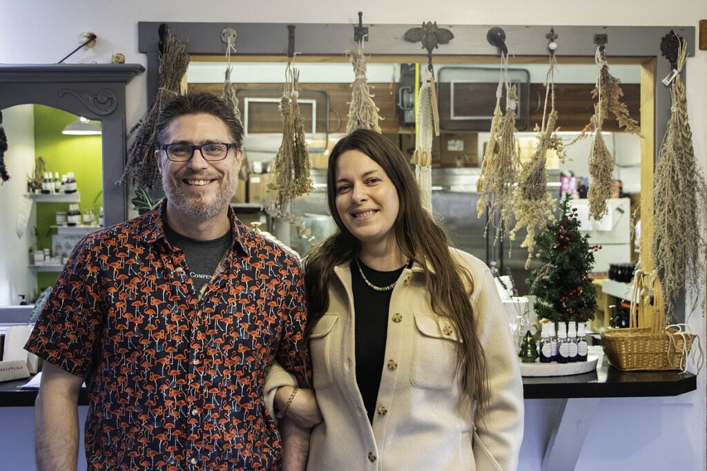 A smiling man and woman stand side by side indoors, in front of a counter decorated with dried herbs and a small Christmas tree—celebrating Spring Success after the Winter Pause. Bottles and jars are displayed behind them.