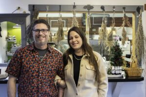 A smiling man and woman stand side by side indoors, in front of a counter decorated with dried herbs and a small Christmas tree—celebrating Spring Success after the Winter Pause. Bottles and jars are displayed behind them.