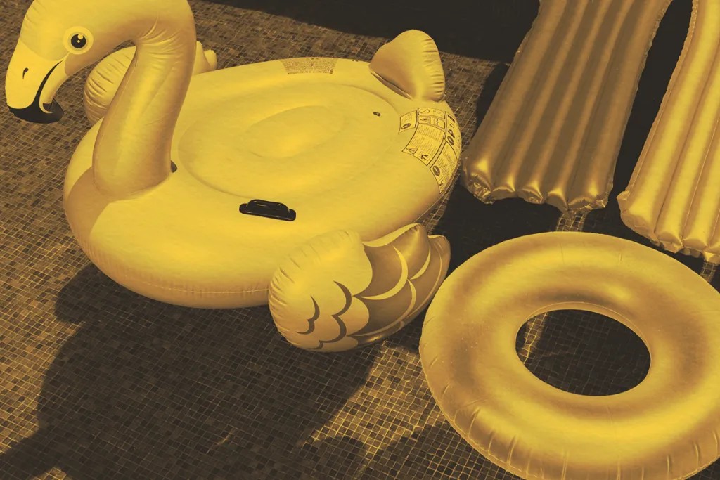 A yellow inflatable swan, an inflatable ring, and an air mattress rest on a tiled poolside surface, evoking Lake Days. The image is enhanced with a yellow-tinted filter for a summery feel.