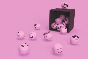 Several white spheres with different smiley faces and emoji expressions sit inside and outside a black box on a pink surface, capturing the playful spirit of Fall in Bigfork and the fun found at September in Bigfork events.