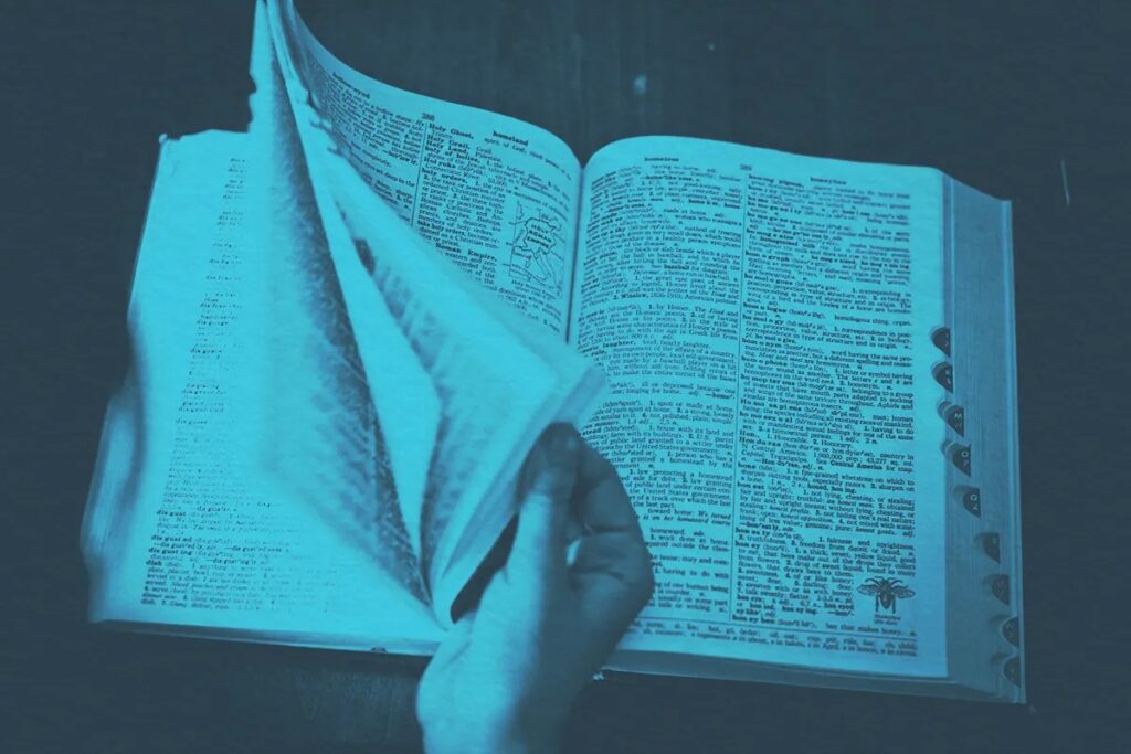 A hand flips through the pages of an open dictionary, with printed text and small illustrations visible on the pages. The image has a blue color overlay, evoking the spirit of Spring and new opportunities for discovery.
