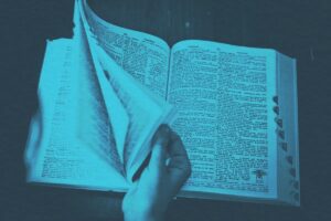 A hand flips through the pages of an open dictionary, with printed text and small illustrations visible on the pages. The image has a blue color overlay, evoking the spirit of Spring and new opportunities for discovery.