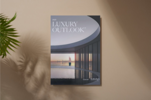 A magazine titled 2025 Luxury Outlook by Glacier Sothebys lies on a beige surface, featuring a modern curved building with large windows overlooking water and a city skyline at sunset. Nearby, a plant casts a shadow—hinting at blooming homes and new beginnings.