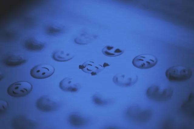 A close-up of various emoji faces displayed on a screen, with a blue tint. The focus is on a smiling face with hearts, capturing the spirit of June joys and new beginnings while other emojis appear blurred in the background.