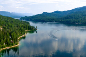 A small boat creates a wake as it travels across the calm, clear waters of Flathead Lake surrounded by dense green forests and distant mountains—a picturesque scene in the heart of Flathead Valley Real Estate.