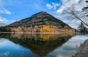 A tree-covered mountain rises beside a calm river, with autumn trees displaying golden leaves. The mountain and trees reflect clearly in the water under a partly cloudy sky—just one of many stunning views near Kalispell Real Estate.