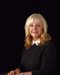 A woman with long blonde hair, wearing a black top with a white collar, sits against a black background. Smiling warmly, she has her hands folded and wears a watch—an approachable face in Flathead Valley Real Estate.