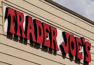 A close-up of a Trader Joe’s store sign with bold red letters outlined in black mounted on a beige, horizontally paneled wall, capturing the community spirit that welcomes shoppers during the cozy season.