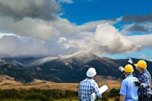 Three men wearing plaid shirts and safety helmets stand in a grassy field, looking and pointing toward distant mountains under a dramatic, cloudy sky. One man holds rolled-up blueprints, planning future listings for summer’s July Journeys.