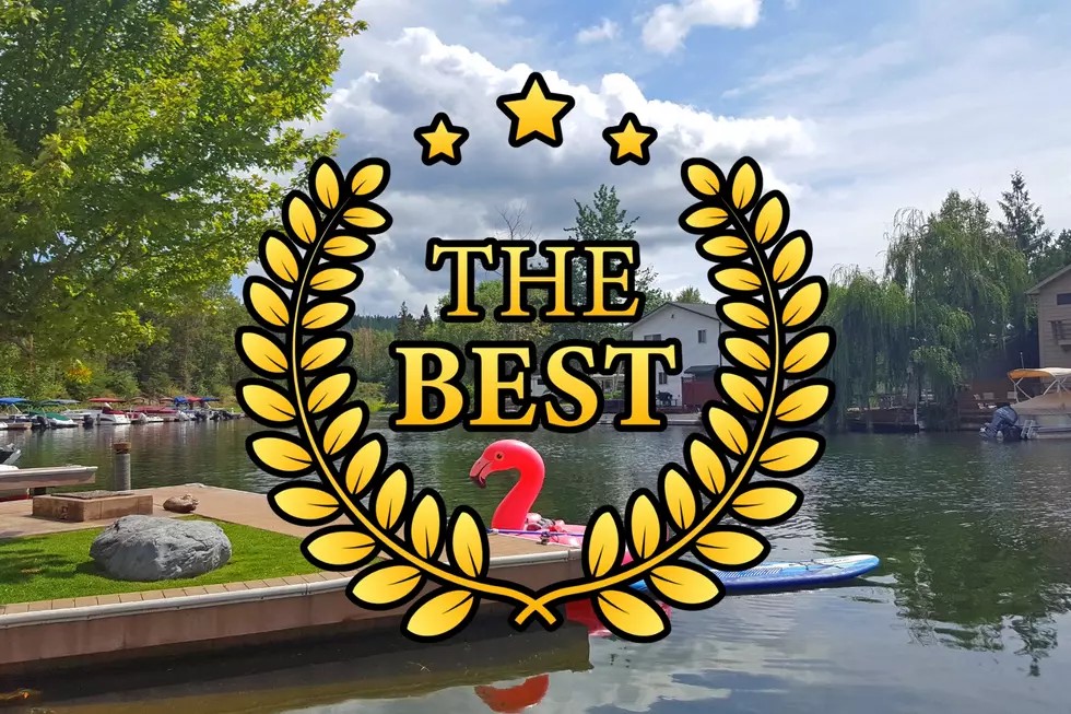 A lakeside scene with docks, boats, and trees under a cloudy sky. A pink flamingo float is on the water, celebrating Bigfork Days. Overlaid is a gold laurel wreath with stars and the words “THE BEST” in bold black letters.