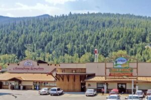 A row of shops, including a restaurant, casino, and gift shop, sits in front of a forested hillside with parked cars and people outside under a partly cloudy sky during September in Bigfork.