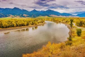 A winding river flows through a valley with autumn-colored trees, surrounded by grassy fields and distant mountains under a cloudy sky—love is in the air as nature’s beauty is heating up the landscape.