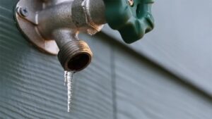 A close-up of an outdoor water spigot with a green handle, mounted on a wall, with a small icicle hanging from the spout—despite freezing temperatures, there's love in the air as winter gives way to a new season.