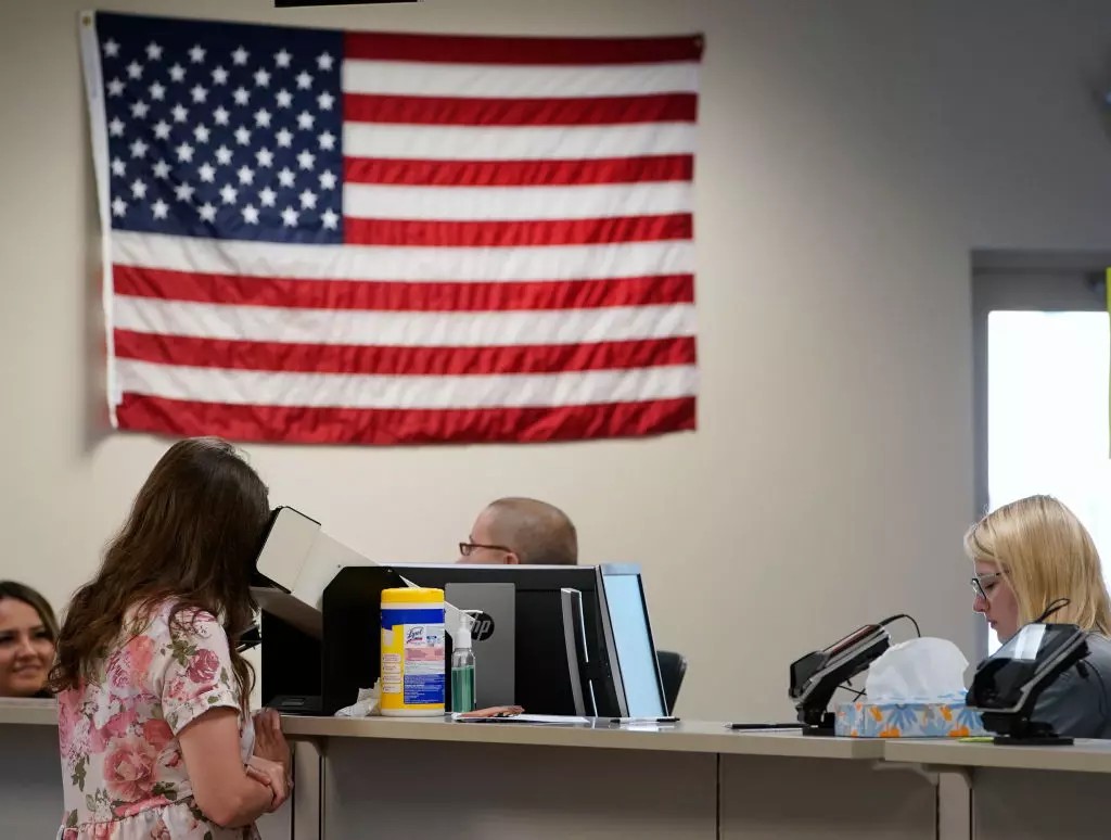 A woman stands at a counter speaking to a staff member in an office with computers, while another staff member works nearby; an American flag hangs on the wall, adding color to the busy scene during the spring season.