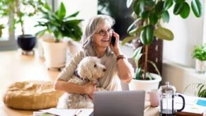 Smiling older woman with gray hair sits at a desk with a laptop, holding a small white dog and talking on the phone, surrounded by green houseplants in a bright room that captures the feel of fall in Bigfork.