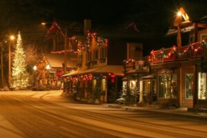 A small town street at night is decorated with festive holiday lights, capturing the cozy season. Buildings glow with colorful string lights, and a large, brightly lit Christmas tree stands at the corner, casting warmth on the snowy road.