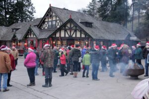 A group of people wearing Santa hats gather outside a festive, Tudor-style building decorated with Community Lights. Some stand near a fire pit while others chat in small groups, surrounded by trees in the background.
