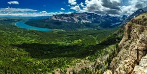 A panoramic view of a lush green valley with dense forests, a turquoise Flathead Lake Home, rugged mountains in the background, and dramatic clouds casting shadows over the landscape. Rocky cliffs appear in the foreground.