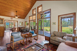 A bright living room with a wood-paneled ceiling, large windows, and glass doors overlooking greenery—this open, airy space embodies the comfort and style often found in Kalispell Real Estate. Cozy furniture and a colorful rug complete the inviting atmosphere.
