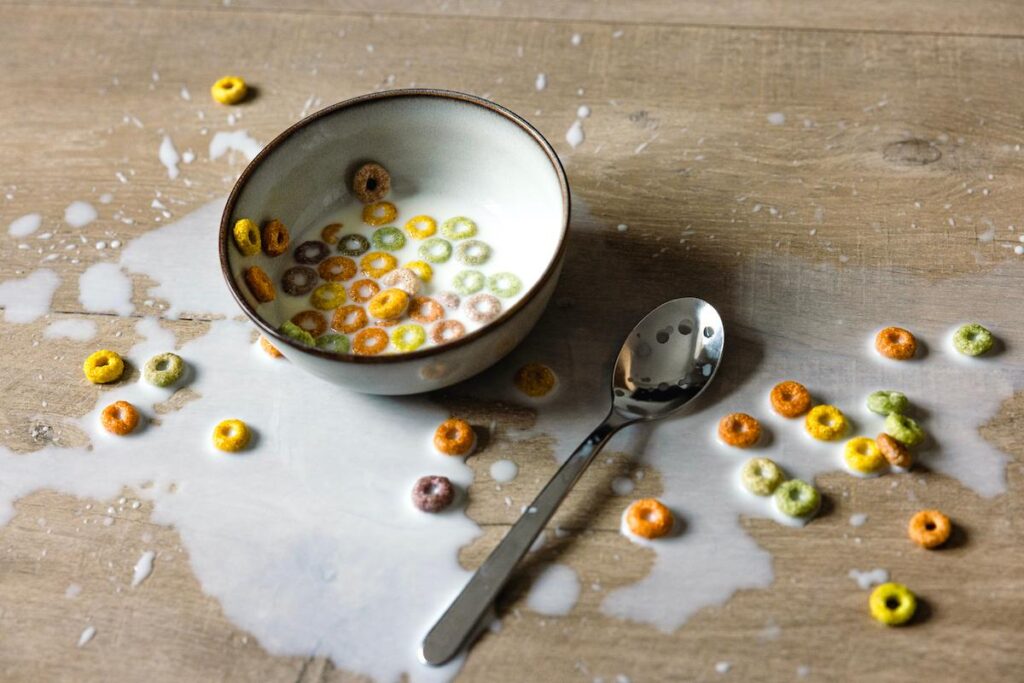 A bowl of colorful cereal with milk sits on a wooden table, surrounded by spilled milk and scattered cereal pieces. A metal spoon rests nearby, capturing a quiet moment in the Season of Action.