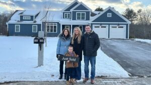 A smiling family of four stands in front of a blue house with snow on the ground. One person holds a Just Sold sign, showing their love for their new home as the market is heating up. Two mailboxes are visible near the driveway.