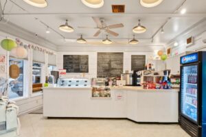 A bright ice cream parlor in Flathead Valley with a white counter, blackboard menus, and a variety of drinks. Decorative paper lanterns hang from the ceiling, and a glass refrigerator stocked with beverages stands on the right.