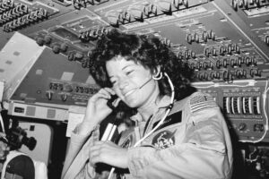 A female astronaut in a NASA flight suit smiles while speaking into a headset microphone inside a spacecraft, surrounded by control panels and switches, embodying the spirit of new beginnings during June celebrations.