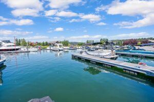 A marina with several boats docked in calm blue water, surrounded by floating docks. In the background are buildings, trees, and a partly cloudy sky.