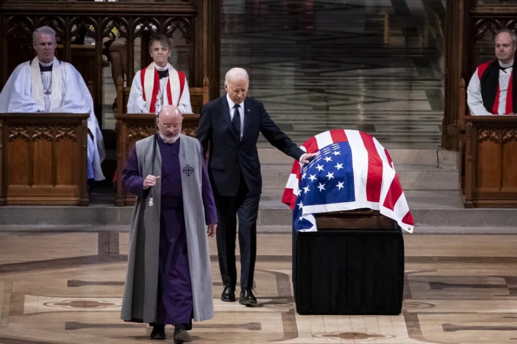 President Joe Biden stands beside a casket draped in an American flag during a church ceremony, as clergy in robes and stoles honor a life marked by Spring Success after a solemn Winter Pause.