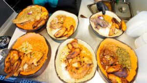 Five plates of food with rice, fried plantains, stewed meats, beans, and vegetables are arranged on a counter, capturing the steady momentum of meal prep as a person in the background serves or prepares the dishes.