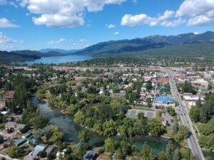 Aerial view of a small town with green trees, tennis courts, and houses by a river, set against mountains and Flathead Lake—a stunning scene ideal for those seeking Flathead Valley real estate or your dream Flathead Lake home.
