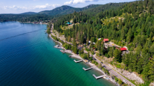 Aerial view of a clear blue lake bordered by a forested shoreline, lakeside homes, small docks, and a winding road, with hills and trees in the background under a partly cloudy sky.