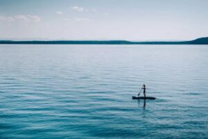 A person stands on a paddleboard in the middle of a calm, expansive body of water under clear February light, with distant tree-lined shores visible on the horizon.