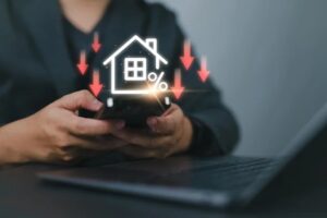 A person using a smartphone near a laptop, with a glowing house icon and downward arrows above the phone, symbolizing movement in mortgage rates or new beginnings amid declining real estate values.