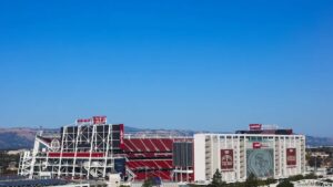 A large football stadium with red seats and a white steel structure, labeled Levi’s Stadium, sits under a clear blue sky with distant hills, embodying the energy of new beginnings and movement.