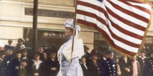 A woman in historical attire holds an American flag upright during a public parade in Flathead Valley, with people and uniformed officers watching near a United Cigar Stores sign, evoking the spirit of a Spring Market celebration.