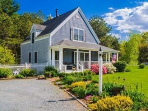 A charming gray-blue house with white trim and a covered front porch welcomes new beginnings, nestled beside a gravel driveway, vibrant flower garden, and white fence—surrounded by green trees under a bright blue sky with scattered clouds.