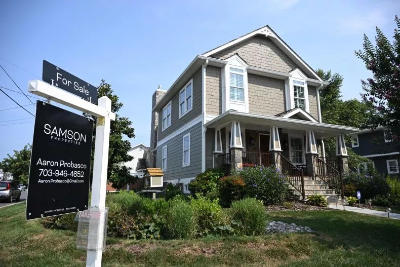 A two-story suburban house with beige siding and white trim sits behind a For Sale sign from Samson Properties on a sunny day. The house, featured in Blooming Homes, has a front porch, green lawn, and landscaped garden.