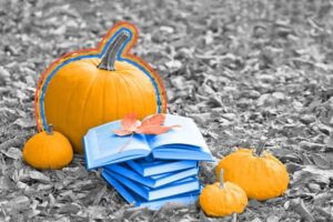 A large pumpkin and three smaller pumpkins sit on fallen leaves next to a stack of blue books, with a red autumn leaf resting on the open book—hinting at folklore tales perfect for sharing within the community.
