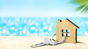 A small wooden house model and a silver key rest on sand with a blurred blue ocean and sky in the background, capturing the essence of Summer Real Estate or a vacation home during the peak Summer Listing Season.