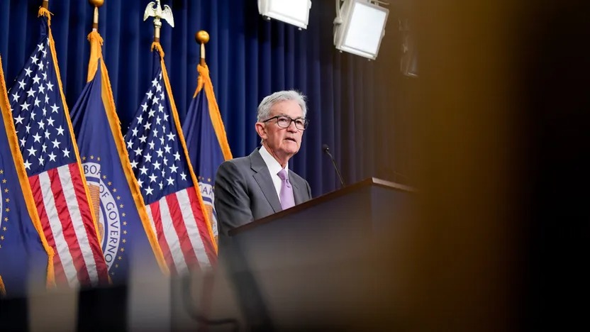 A man in a suit and tie stands at a podium, speaking, with American flags and Full Federal Reserve emblems behind him under bright lights—capturing the energy of an important August event.