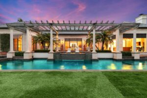 Luxurious modern house with large windows, palm trees, and a spacious swimming pool in front, glowing with August Reflections at sunset. A pergola sits above the pool as warm lights highlight the manicured green lawn in this Season of Action.