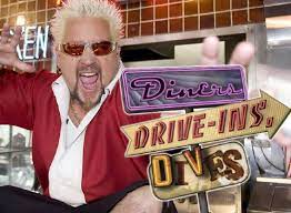 A man with spiked blonde hair and sunglasses, wearing a red and white jacket, poses enthusiastically next to a colorful Diners, Drive-Ins, Dives sign inside a retro-style diner—spreading Merry Christmas cheer for a Bright New Year.