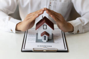 A person’s hands form a protective gesture over a model house placed on an insurance document, evoking the need for security during the changing season and cool crisp days of fall.