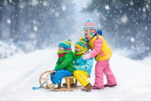 Three children dressed in colorful winter clothes play in the snow; two sit on a wooden sled while one stands behind, ready to push. Amid this Winter Pause, snow falls heavily around them in a snowy forest setting.