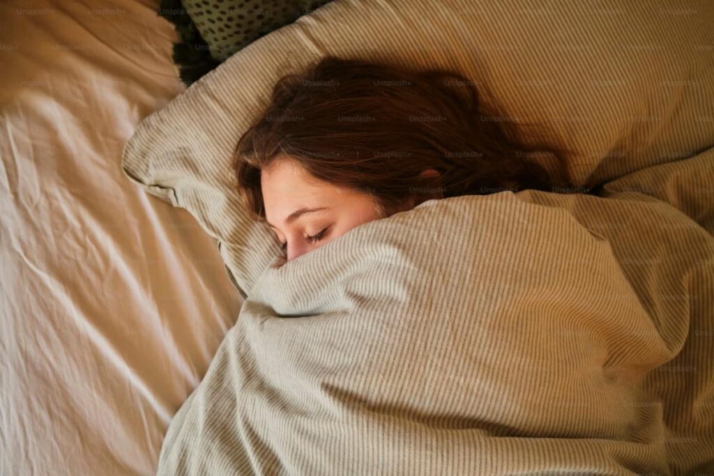 A person with brown hair sleeps on their side in bed, mostly covered by a beige striped blanket, with only part of their face visible above the covers—resting peacefully as spring brings new opportunities.
