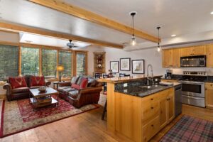 Open-concept living room and kitchen with wood cabinets, stainless steel appliances, granite countertops, leather sofas, tartan rug, and large windows letting in natural light—ideal for buyers seeking comfort and style in the sellers process.