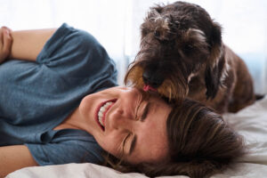 A woman lying on a bed laughs as a scruffy dog licks her head. Sunlight shines through the window, filling the room with a winter glow and creating a bright, playful atmosphere of warm wishes.