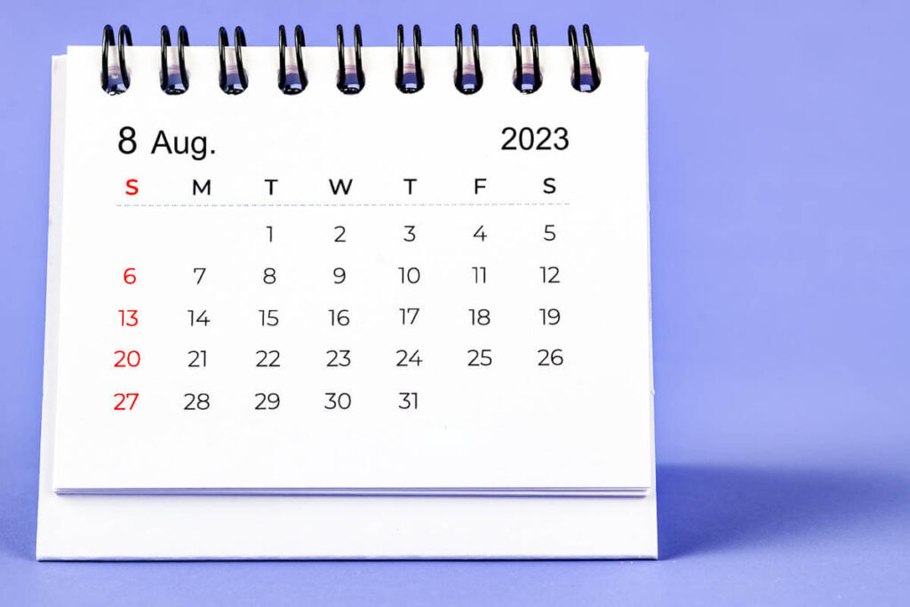 A small spiral-bound desk calendar displays the full month of August 2023 on a purple background, with dates arranged in a grid and Sundays marked in red.