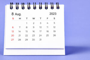 A small spiral-bound desk calendar displays the full month of August 2023 on a purple background, with dates arranged in a grid and Sundays marked in red.